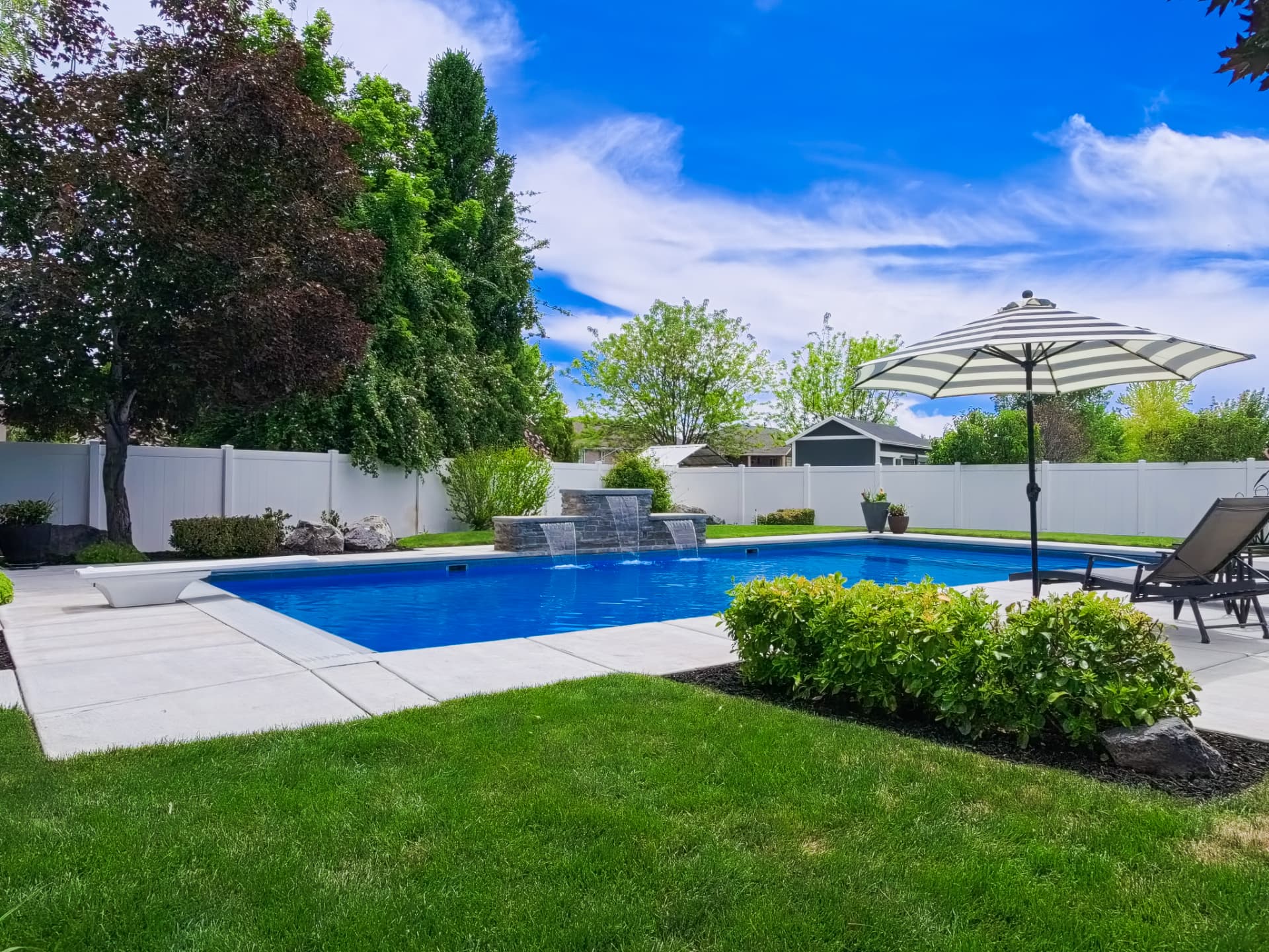 Rectangle vinyl inground swimming pool with stacked-stone waterfall, striped patio umbrella and manicured landscaping in a Kaysville, Utah backyard by Barr Pool Builders