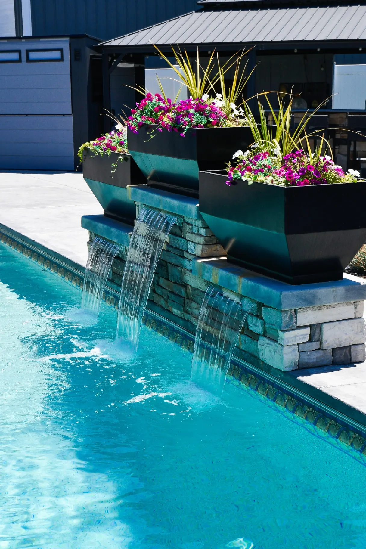 Custom vinyl swimming pool with tiered stone waterfall feature and floral planters in a Syracuse, Utah backyard by Barr Pool Builders