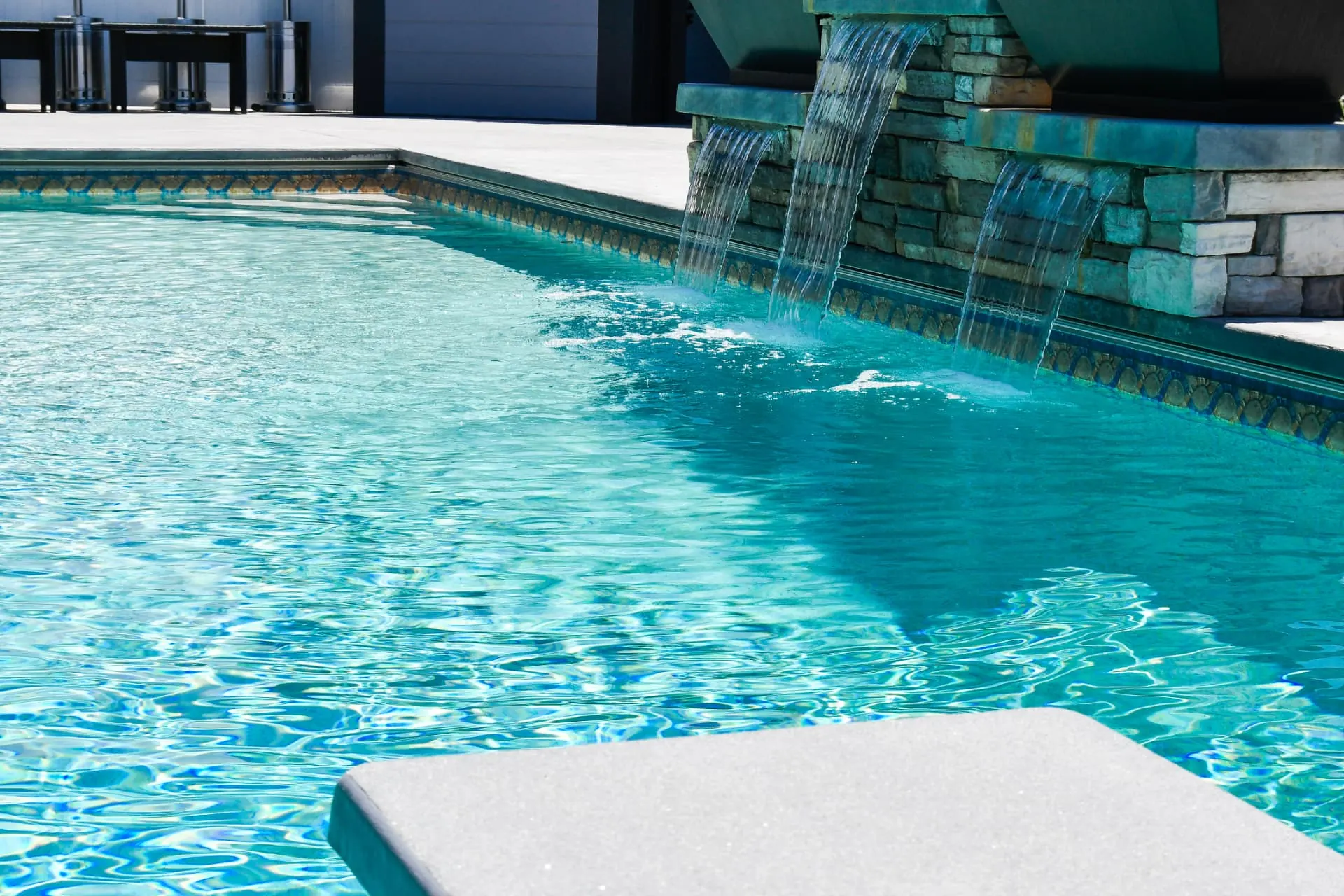 Close-up of stone waterfall feature cascading into a custom vinyl swimming pool with mosaic tile trim and diving board in Layton, Utah by Barr Pool Builders