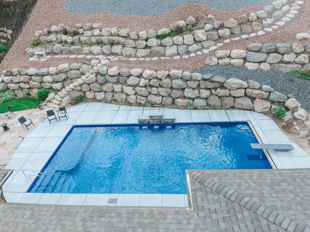 Aerial drone view of a custom rectangle vinyl swimming pool with diving board, stone patio and terraced boulder retaining wall in Layton, Utah by Barr Pool Builders