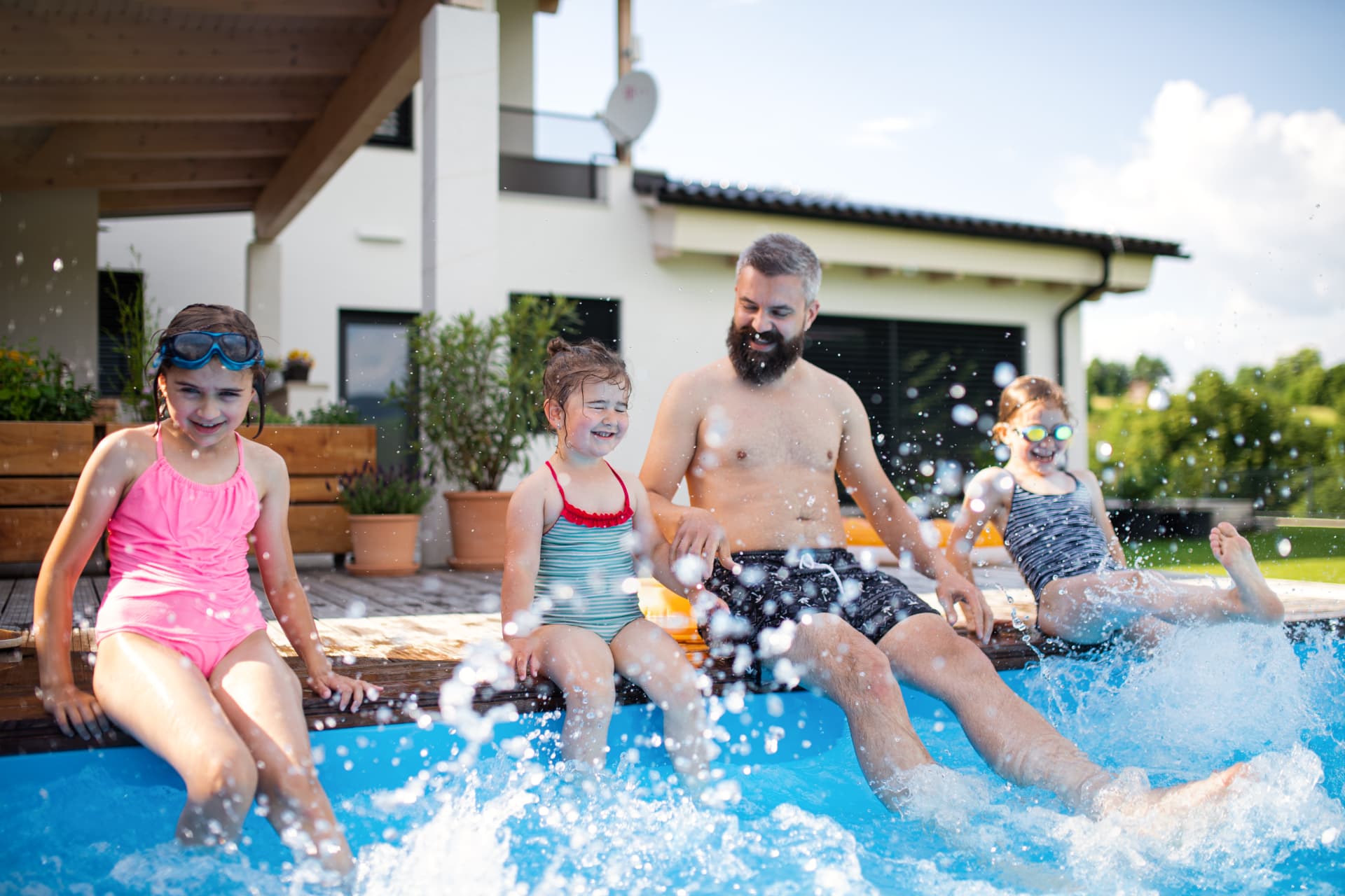Father and three young daughters laughing and splashing at the edge of a backyard swimming pool built by Barr Pool Builders in Northern Utah